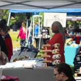 Black woman dressed in red smiling at two women looking at her handmade items at Mavuno