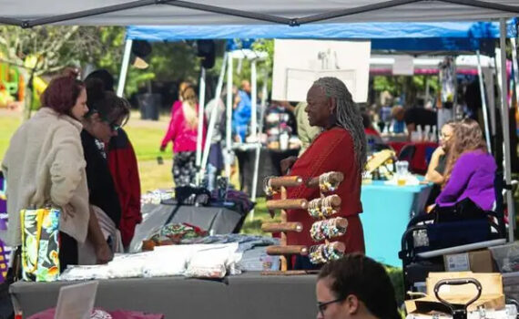 Black woman dressed in red smiling at two women looking at her handmade items at Mavuno