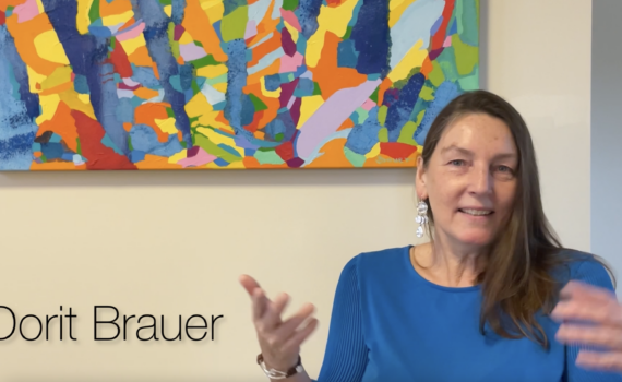 Woman with long dark hair in a blue top speaks in front of multicolored painting