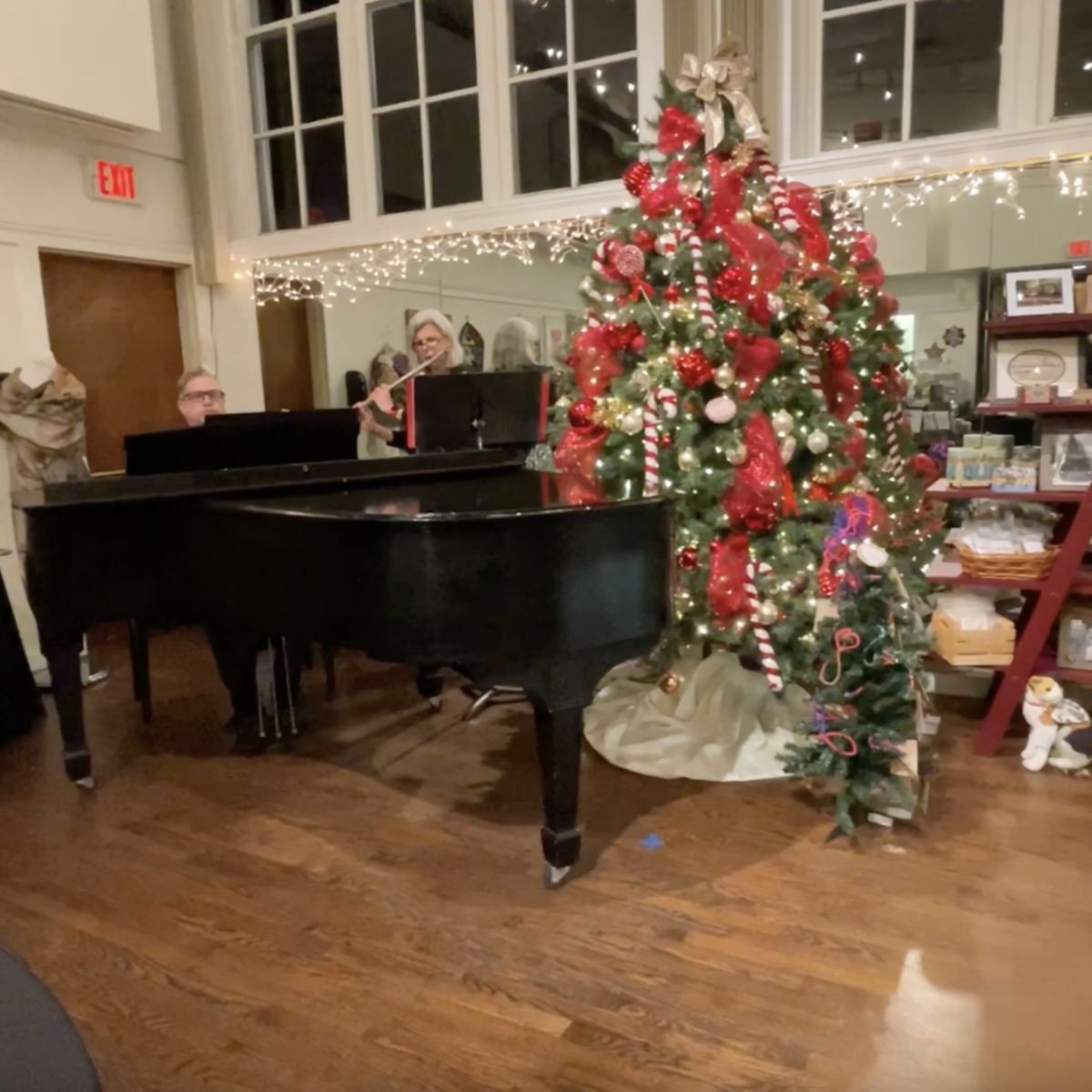 A woman plays the flute with a man at the piano next to a Christmas tree with red ribbons and lights