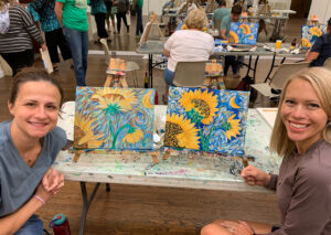 Two woman smiling seated beside the paintings they have created